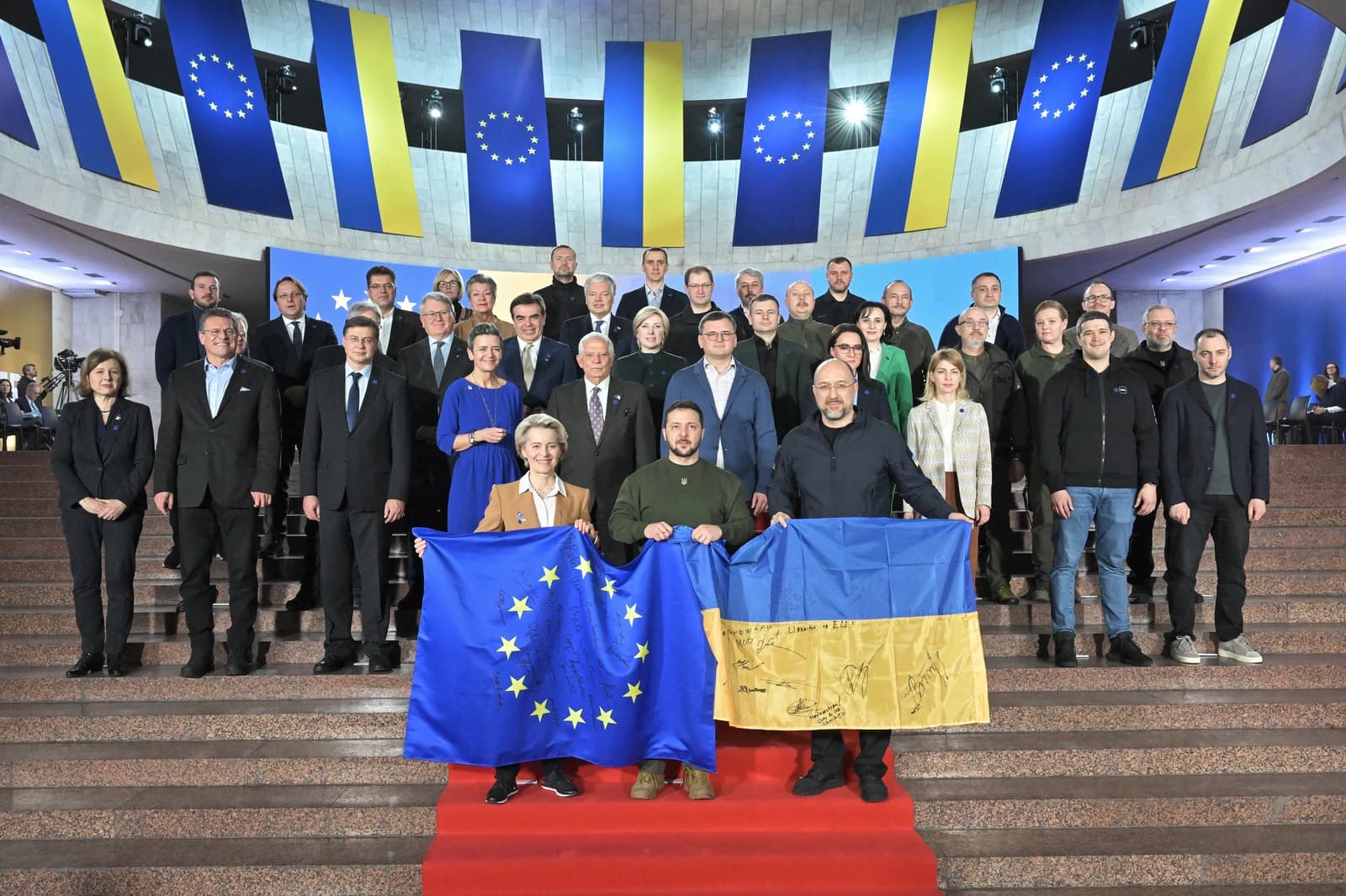 Ursula von der Leyen, Volodymyr Zelensky, and Denys Shmyhal hold flags with other officials.