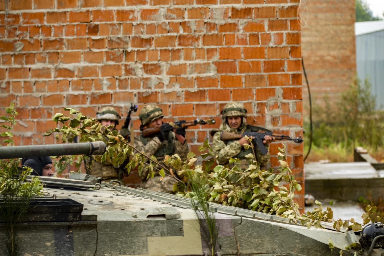 Ukrainian soldiers in camouflage uniforms and helmets, with automatic rifles, near an armored vehicle and brick wall.