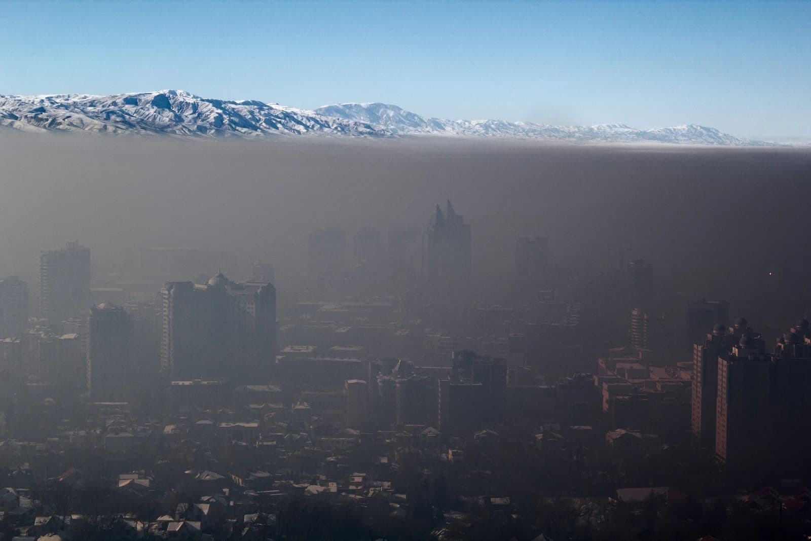 Almaty cityscape shrouded in thick smog, with snow-capped mountains visible above the haze.