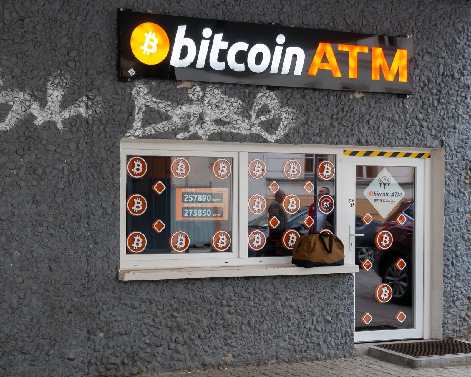 Bitcoin ATM sign above a window with cryptocurrency stickers and exchange rates.
