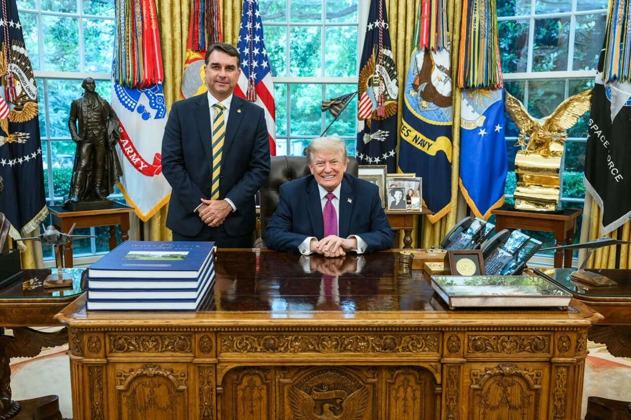 Donald Trump, former U.S. President, sits at a desk with Flávio Bolsonaro standing beside him.