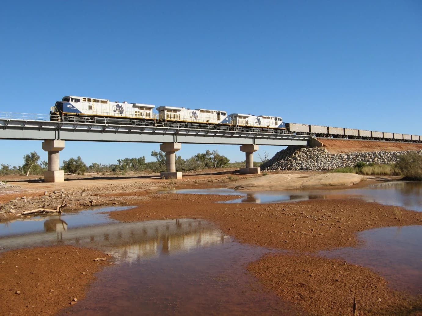 FMG Iron Ore Train, with multiple locomotives, crosses a bridge over a reddish-brown riverbed.