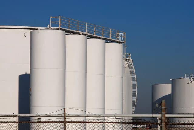 Several large white refinery storage tanks with ladders and railings under a blue sky.