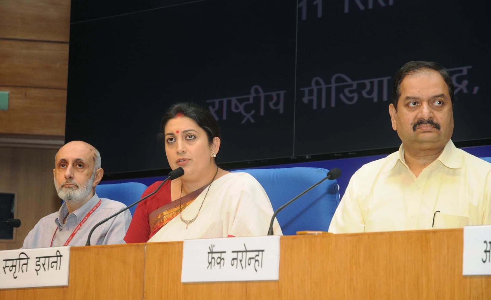Smriti Irani speaking at a press conference in New Delhi, flanked by two men.