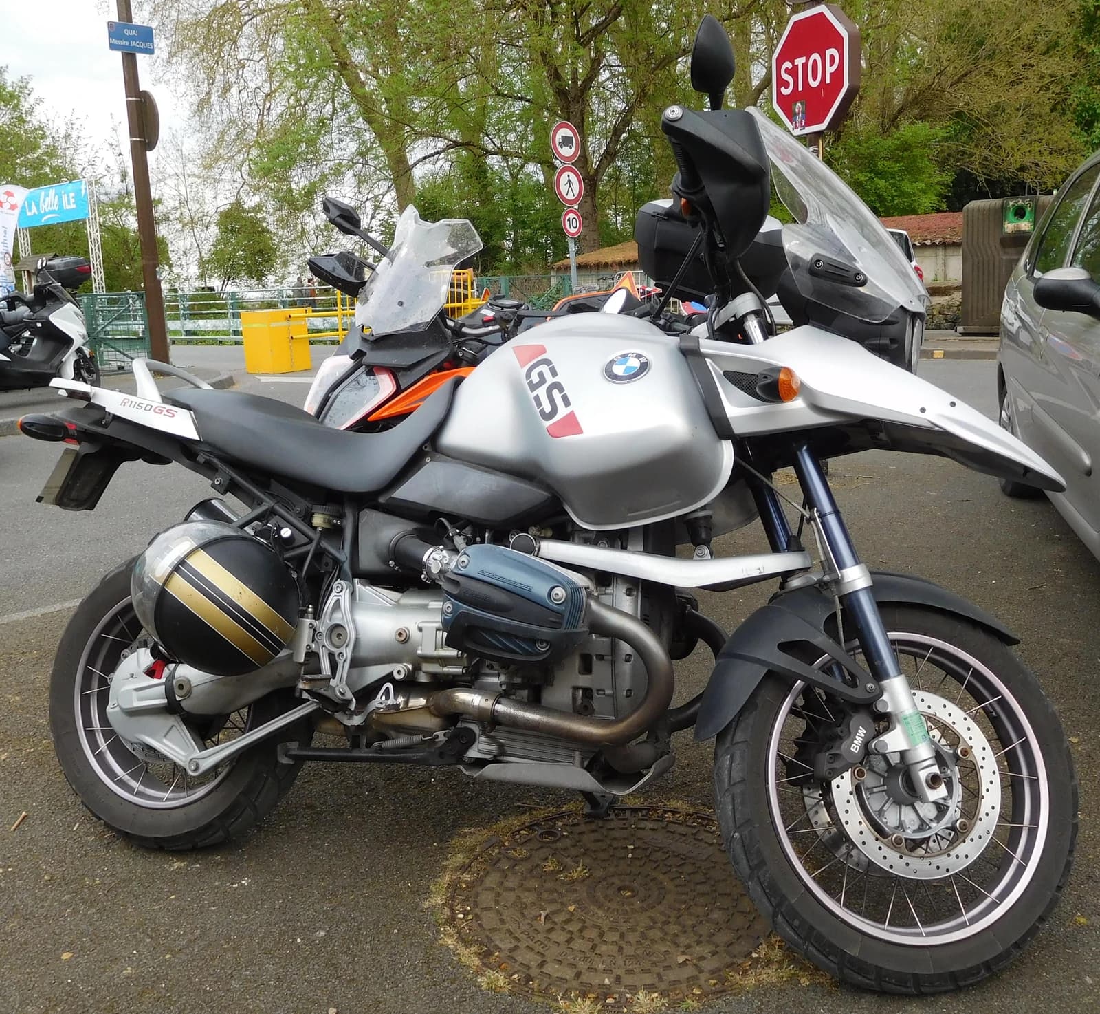 Silver BMW R 1150 GS motorcycle with a black and gold helmet attached, parked on a street.