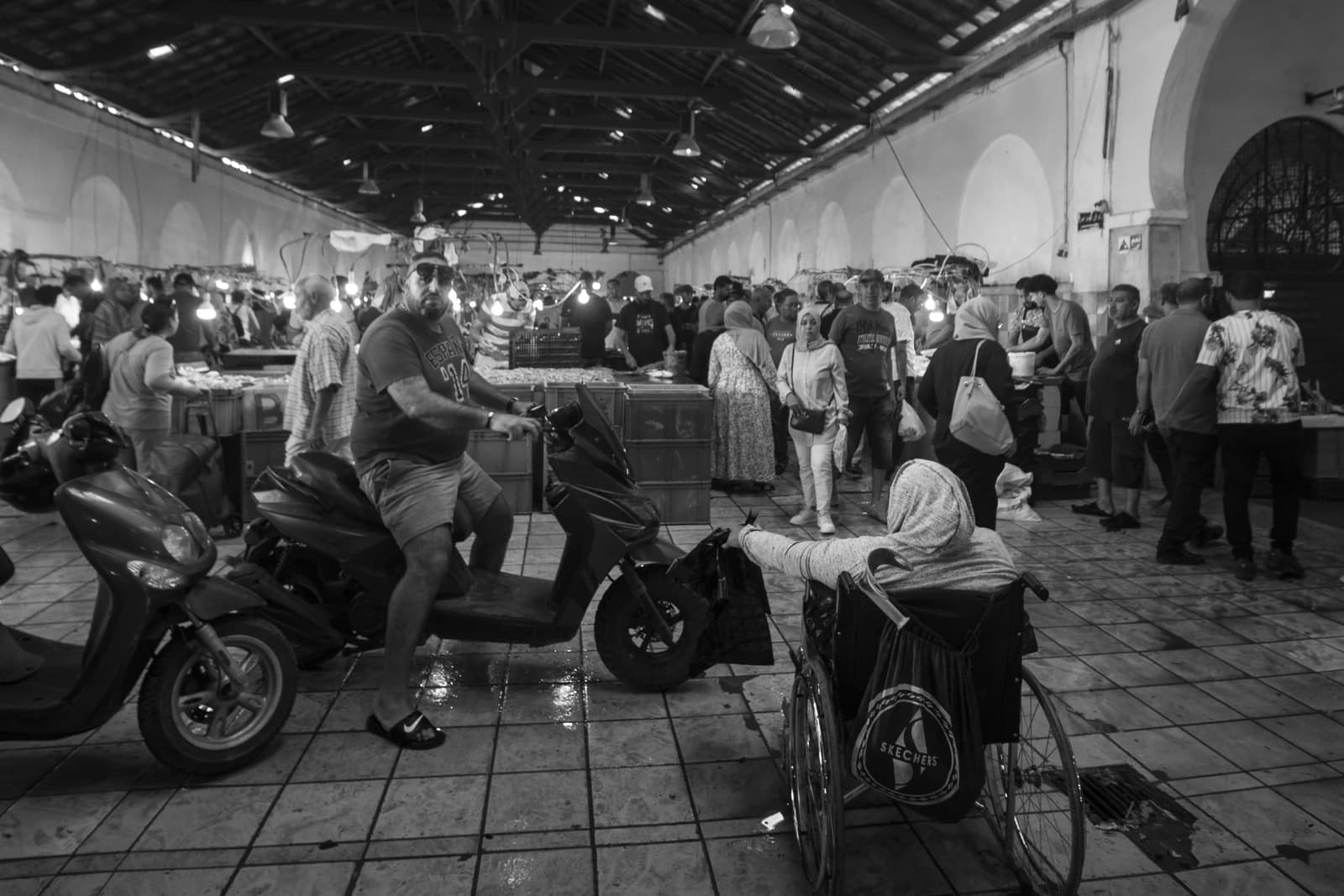 People gathered inside a market building, some standing, one person on a scooter, another in a wheelchair.