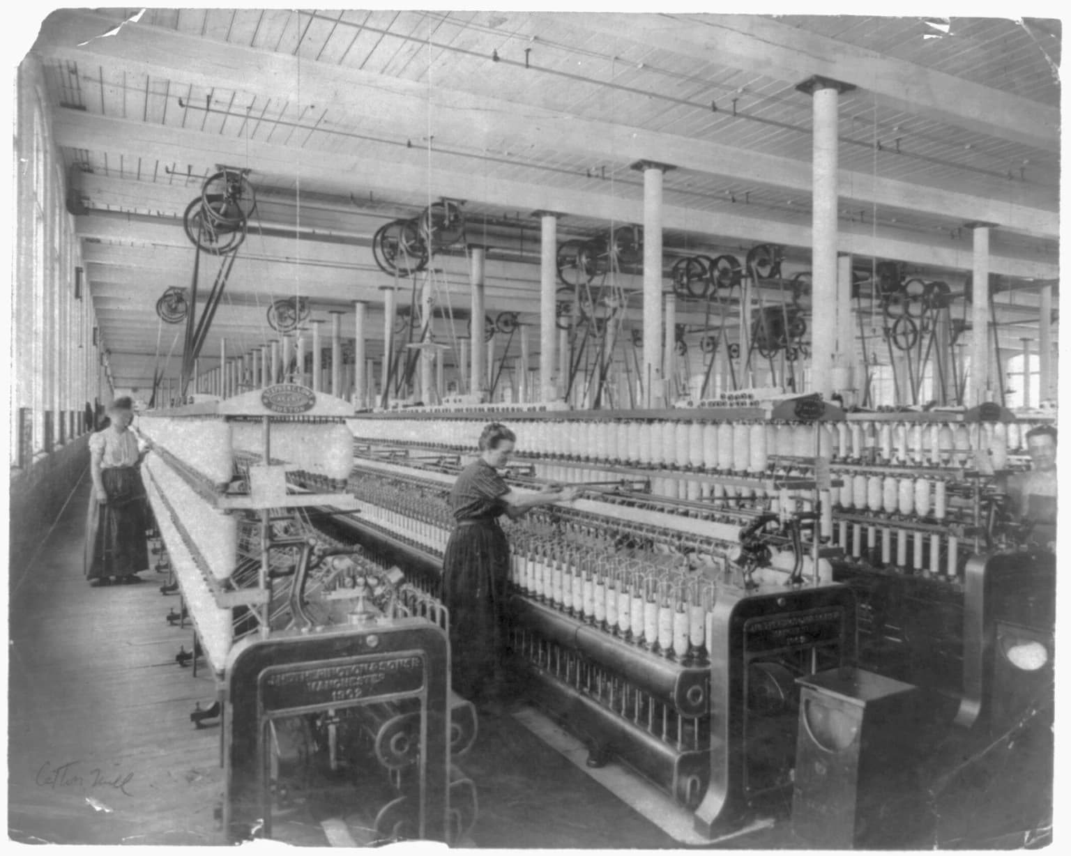 Two women operating textile machinery inside a large cotton mill.