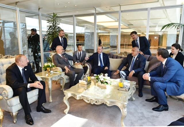 Multiple leaders in suits seated around ornate tables in a formal meeting room.