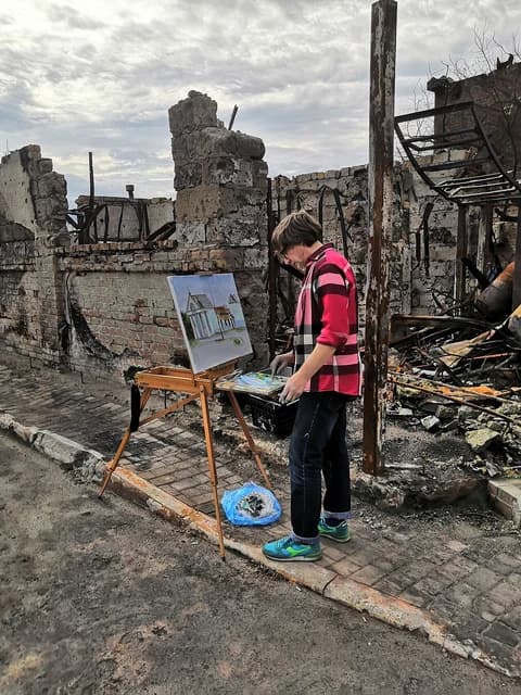 An artist paints a landscape on an easel in front of a destroyed building.