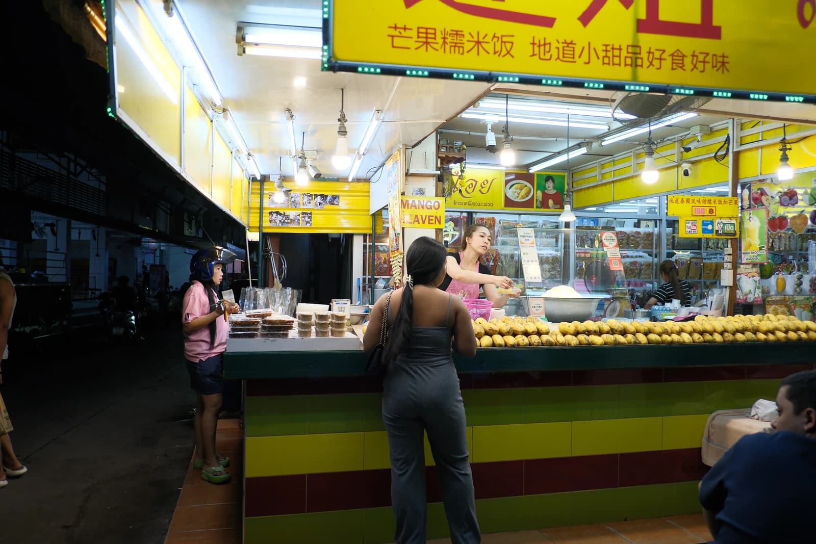 Customers order fresh mango treats from a vendor at a brightly lit street food stall.