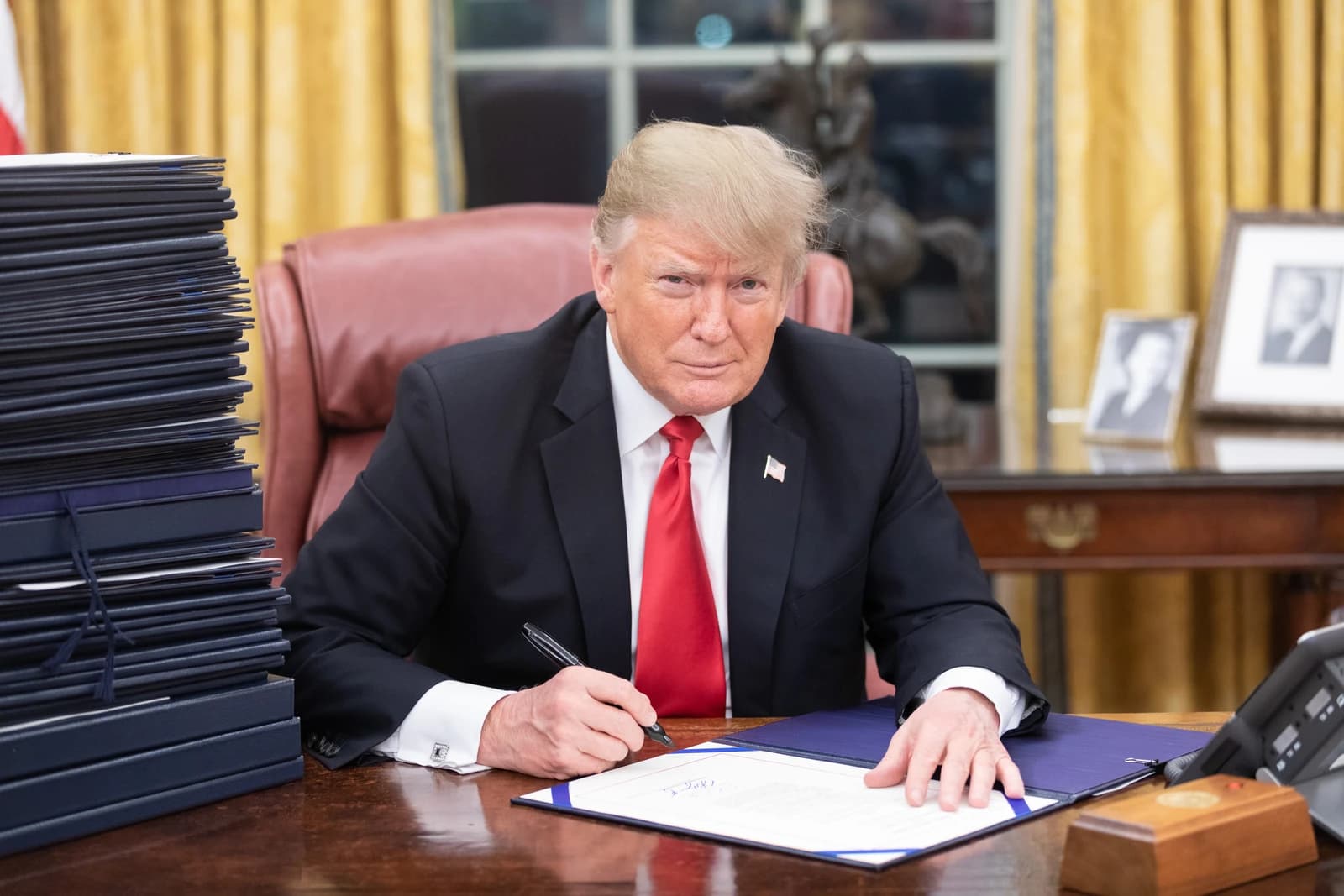 President Donald Trump seated at his desk, signing a document in the Oval Office.