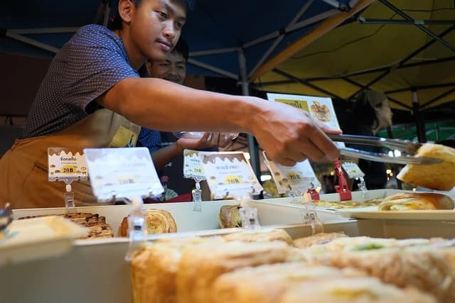 Vendor uses tongs to pick up a baked pastry from a display at an outdoor market.
