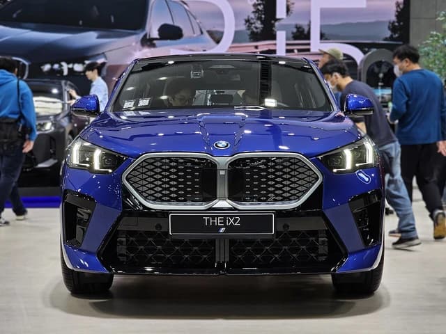 Blue BMW iX2 electric vehicle displayed indoors, with people in the background.
