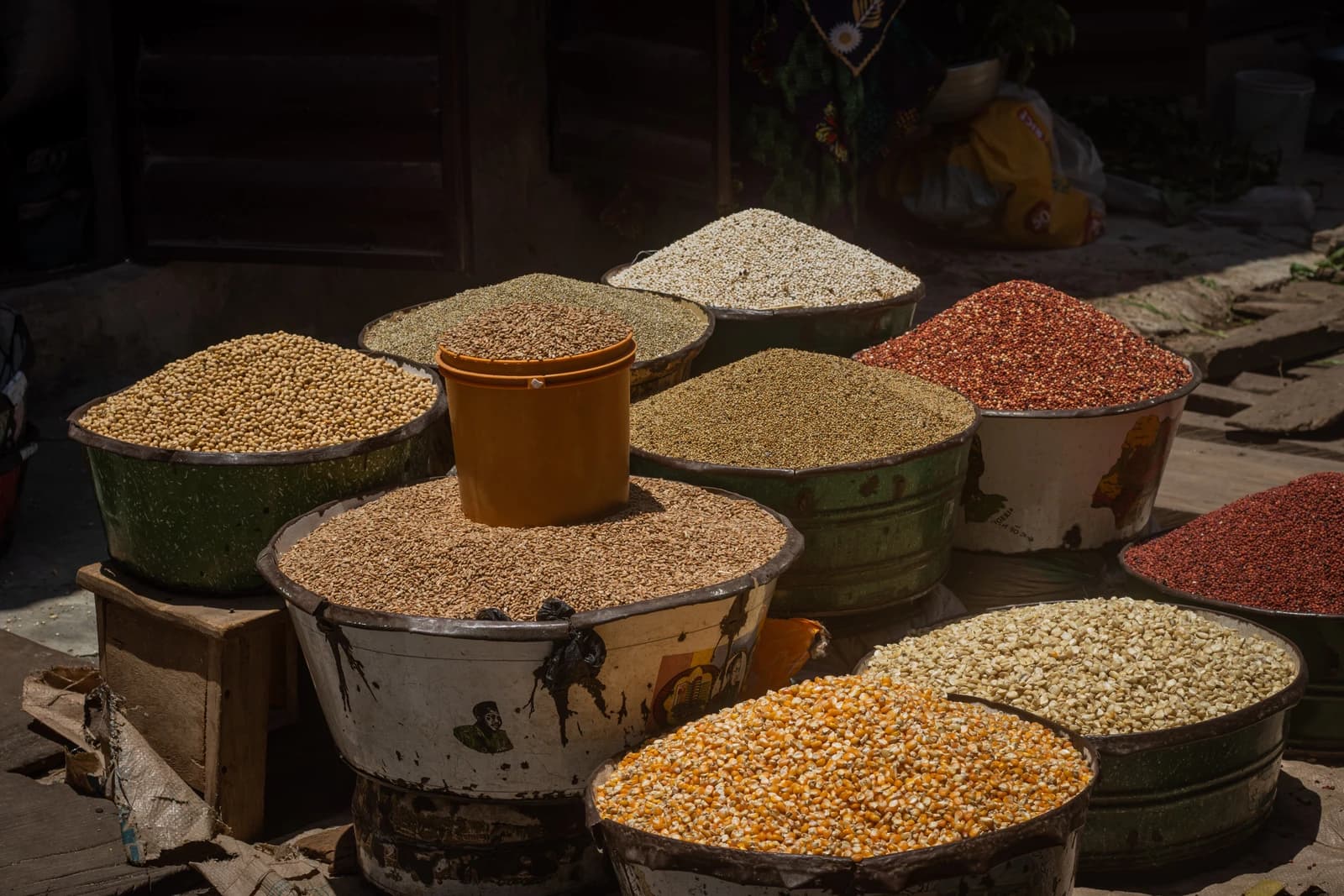 Different varieties of seeds, including maize, piled high in large basins at an outdoor market.