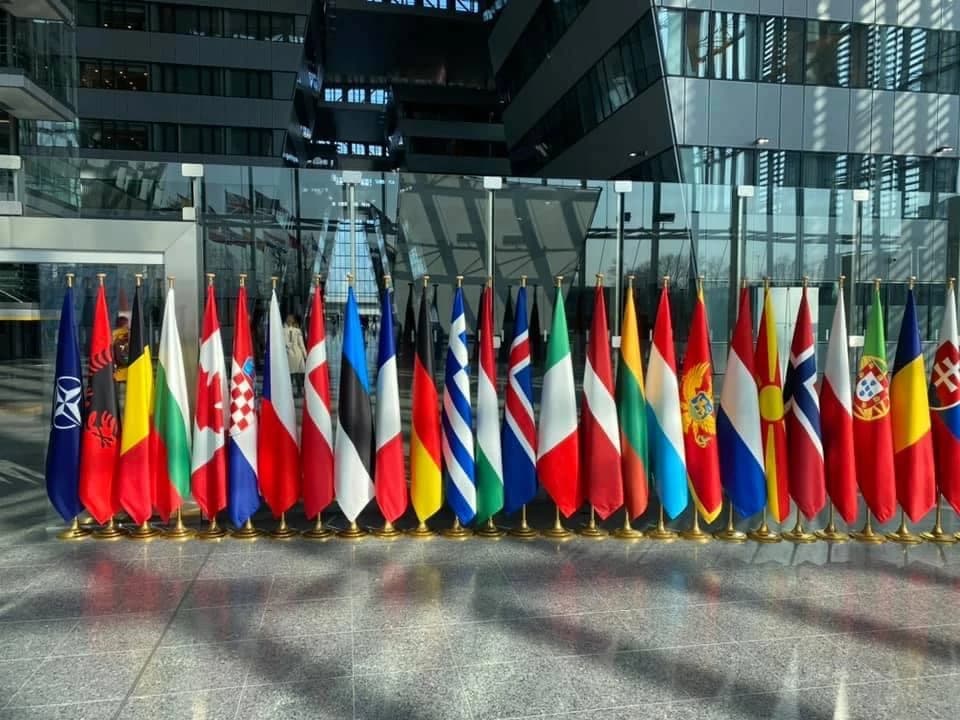 Flags of NATO member nations standing in a row inside a modern building.