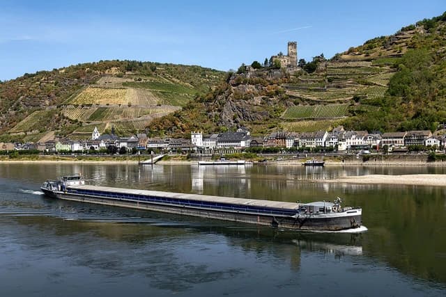 A long barge sails on a wide river with exposed sandbanks and a town on the bank.