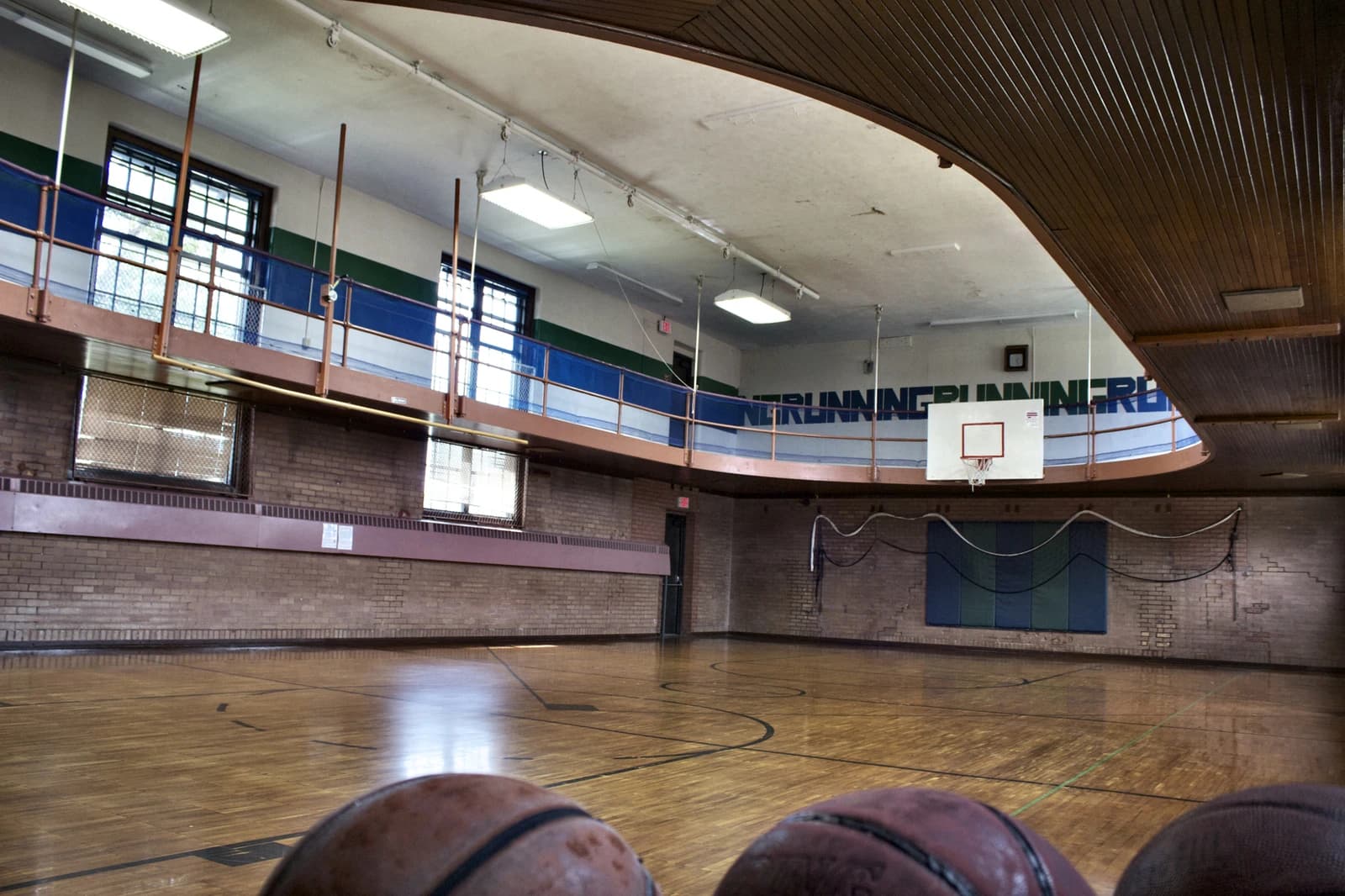 Interior of a school gymnasium with a hardwood floor, basketball hoop, and an elevated running track.