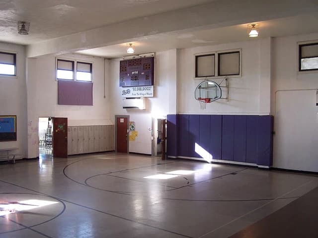 An empty, dated school gymnasium with a scoreboard and basketball hoop.