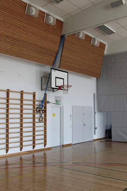 An empty gymnasium with wooden floors, climbing wall bars, and a basketball hoop.