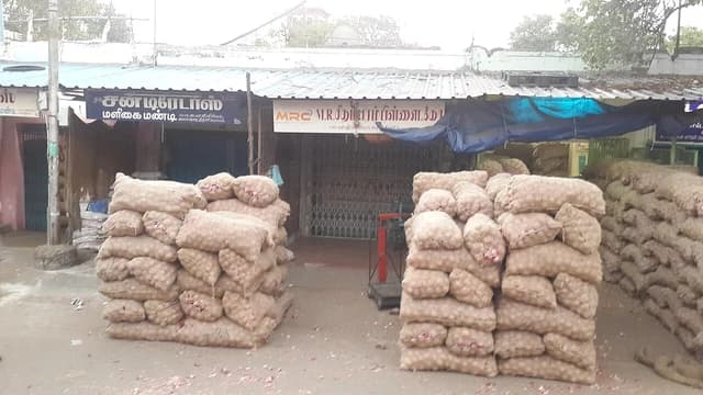 Stacks of mesh sacks filled with onions on the ground in front of market stalls.