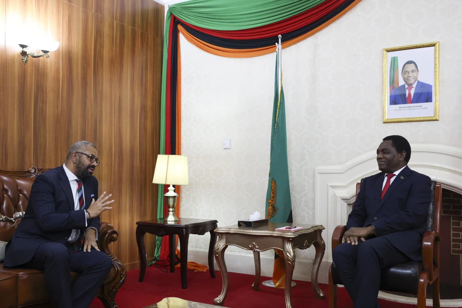 President Hakainde Hichilema of Zambia sitting in a chair, facing left, with a portrait behind him.