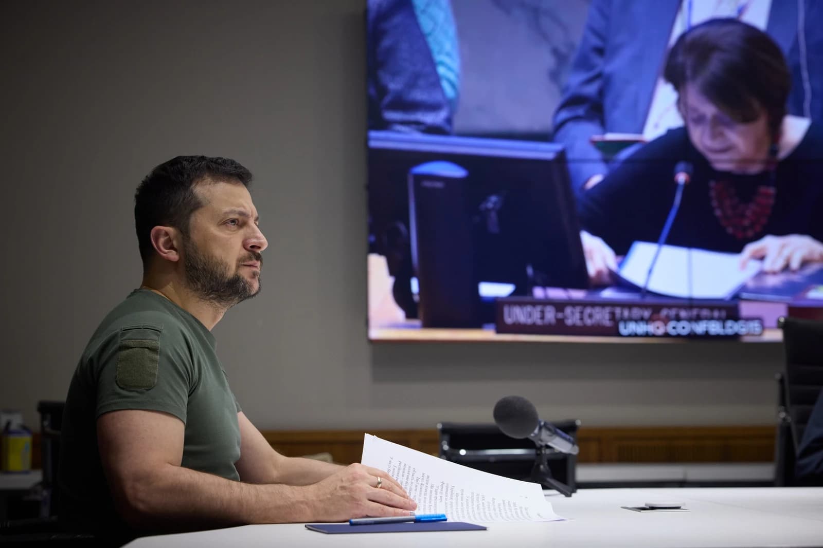 Volodymyr Zelenskyy, President of Ukraine, seated at a desk with papers and a microphone.