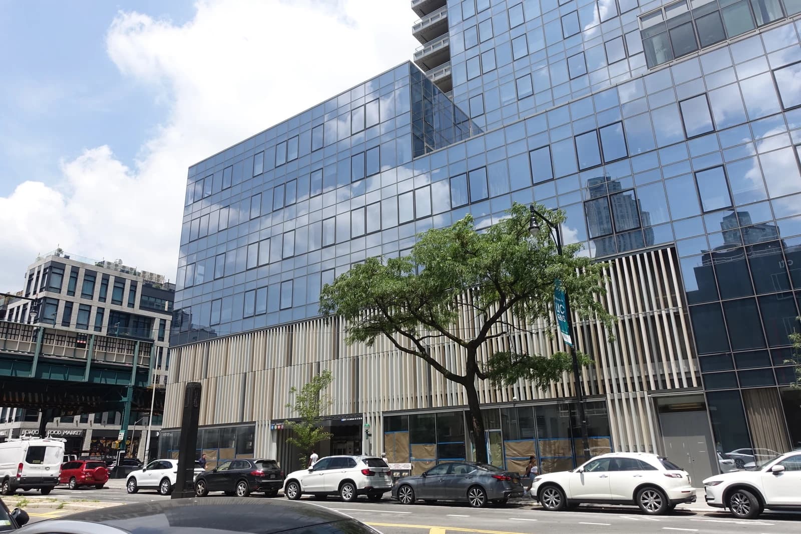 Modern glass skyscraper with parked cars and elevated subway line in an urban setting.