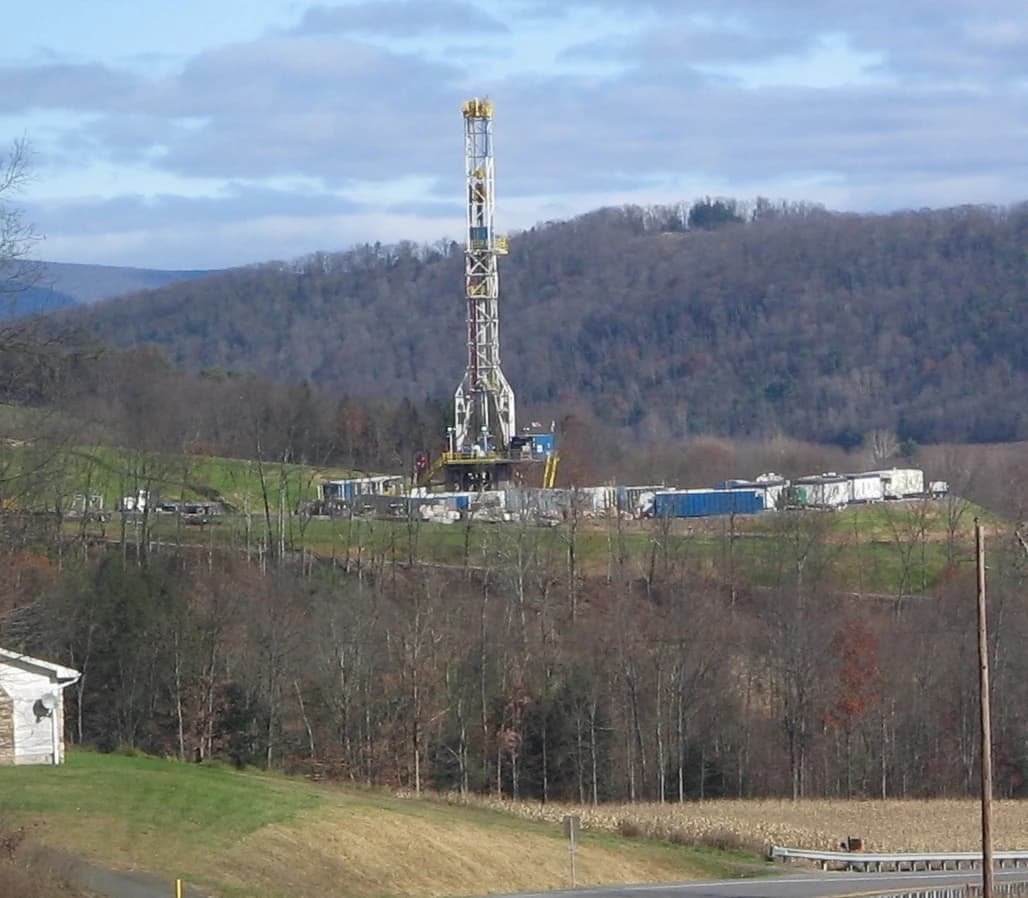 Natural gas drilling tower operates on a hillside in a rural, wooded landscape.