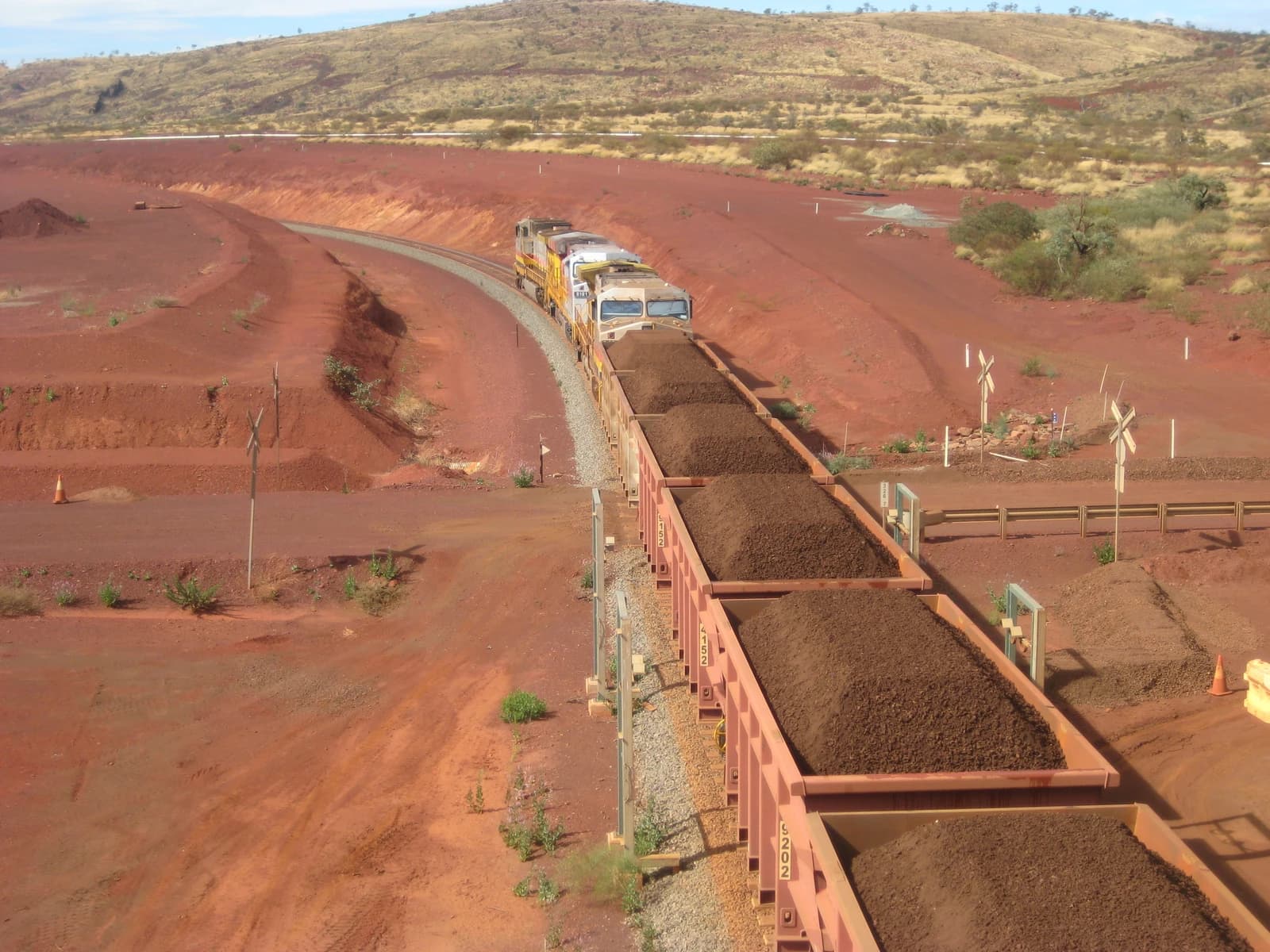 An iron ore train with multiple full carriages moves along a track in a reddish-brown landscape.