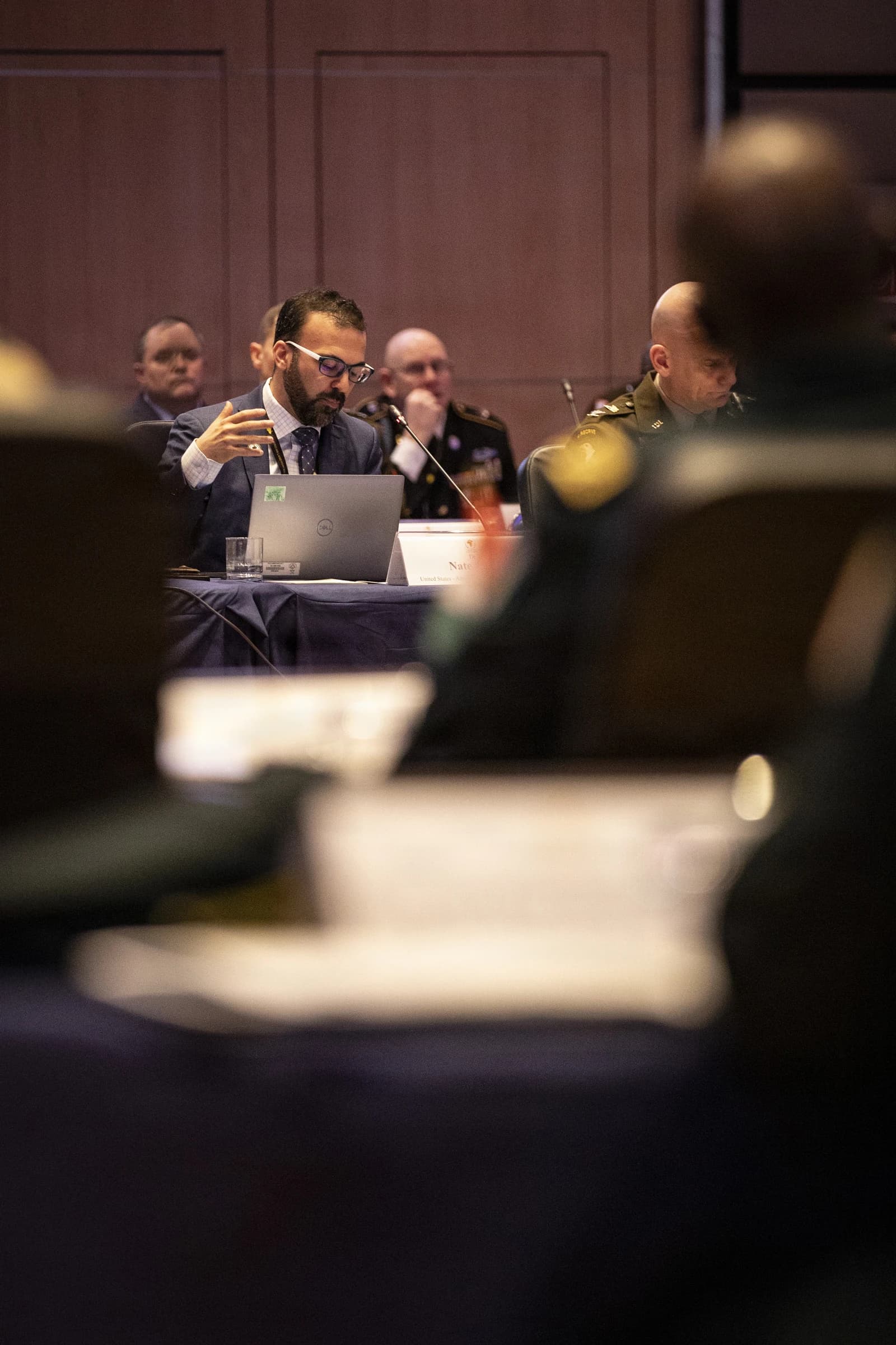A man in a suit speaks at a conference table with a laptop, other panelists in background.