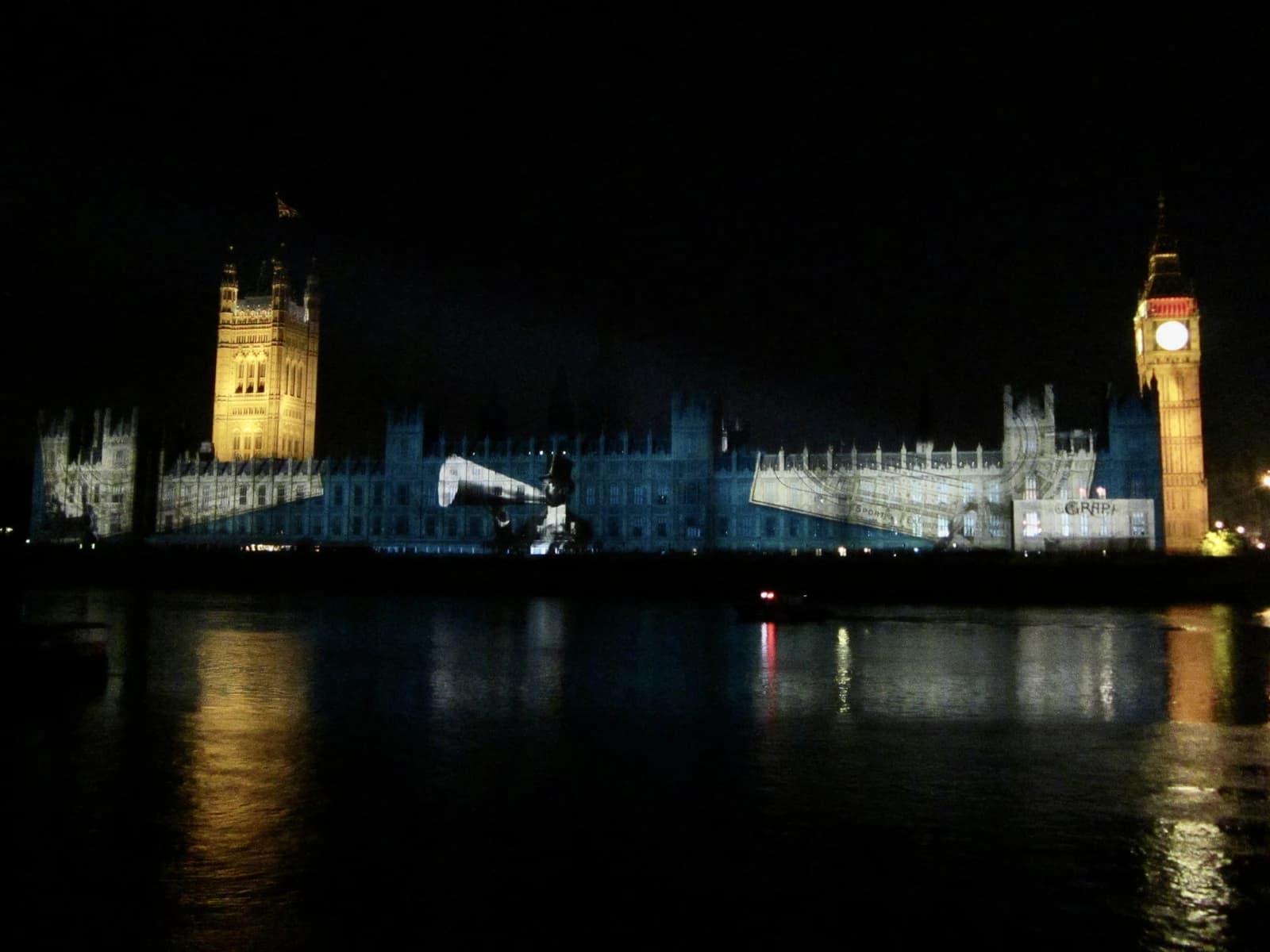 The Houses of Parliament and Big Ben illuminated at night by projection, with reflections on the Thames.
