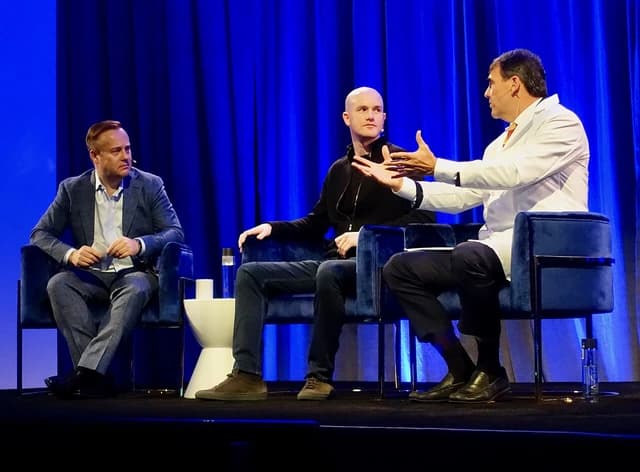 Three men on a stage, one gesturing while speaking, two others listening.