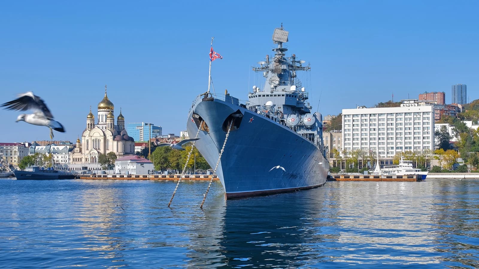 Russian Varyag cruiser docked at the Port of Vladivostok with city buildings and a golden-domed church in the background.