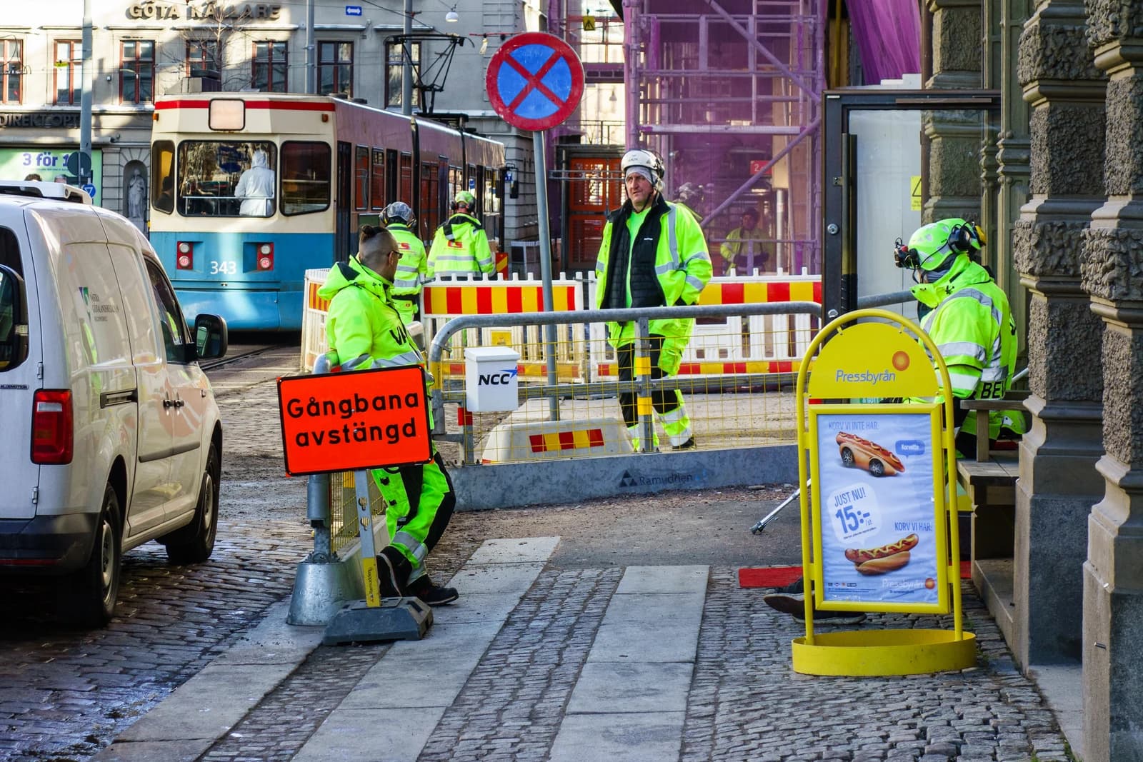 Construction workers in high-visibility jackets and helmets stand on a cobblestone street at a work site.