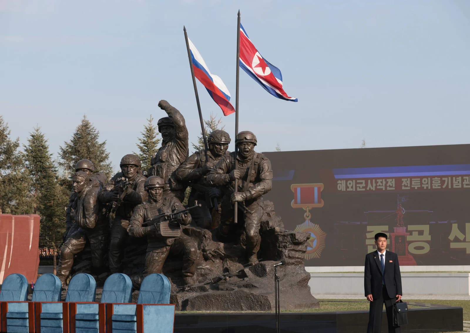 An official stands near a military monument with North Korean and Russian flags flying.