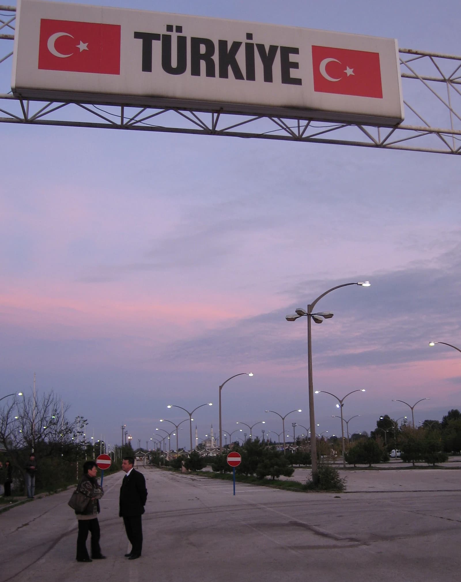 Overhead sign at Kapikule border crossing into Turkey, with two people standing on the road.