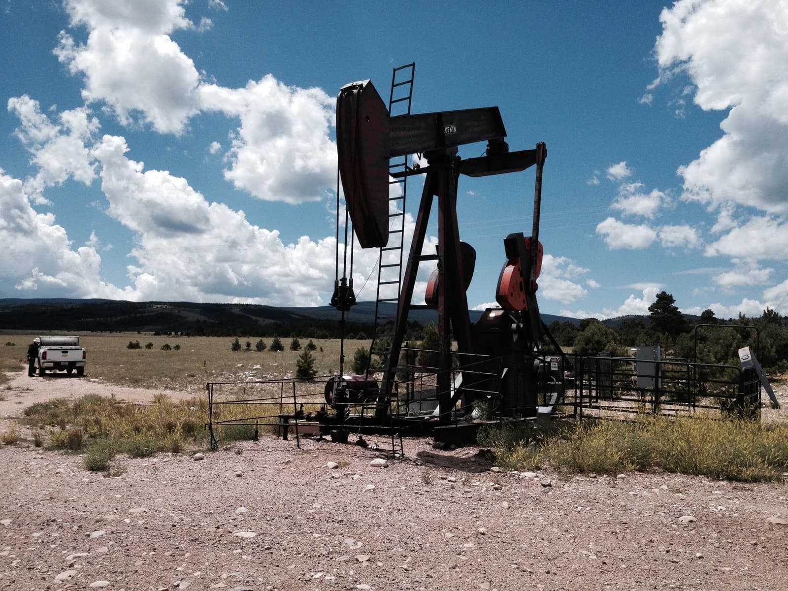 An oil pumpjack stands in a rural landscape, with a pickup truck in the distance.