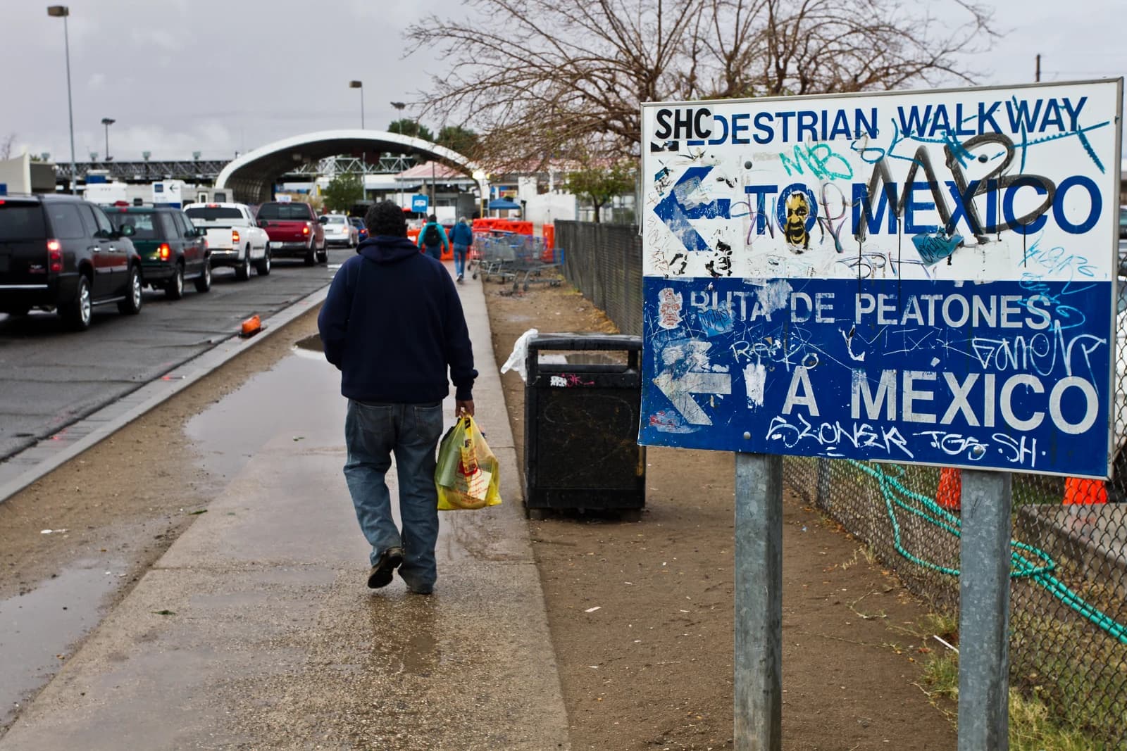Man carrying bags, walking on a sidewalk toward a pedestrian border crossing to Mexico.