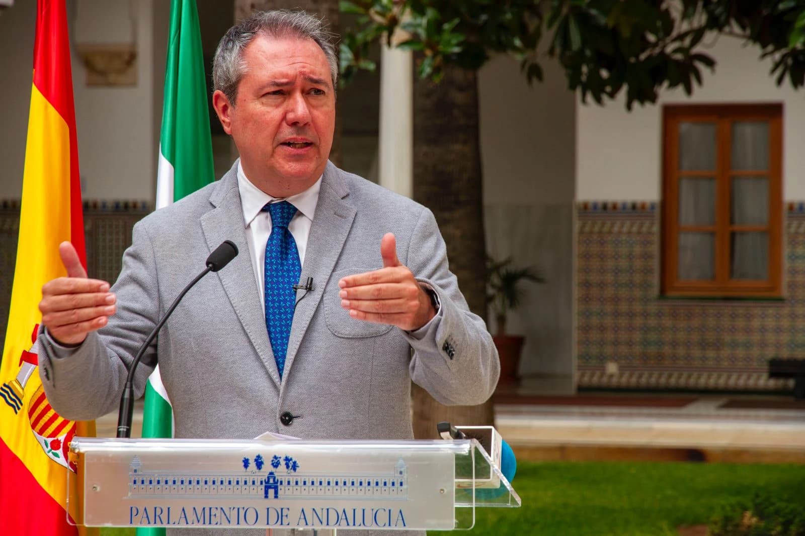 Juan Espadas Cejas, speaking at a podium with flags in the background.