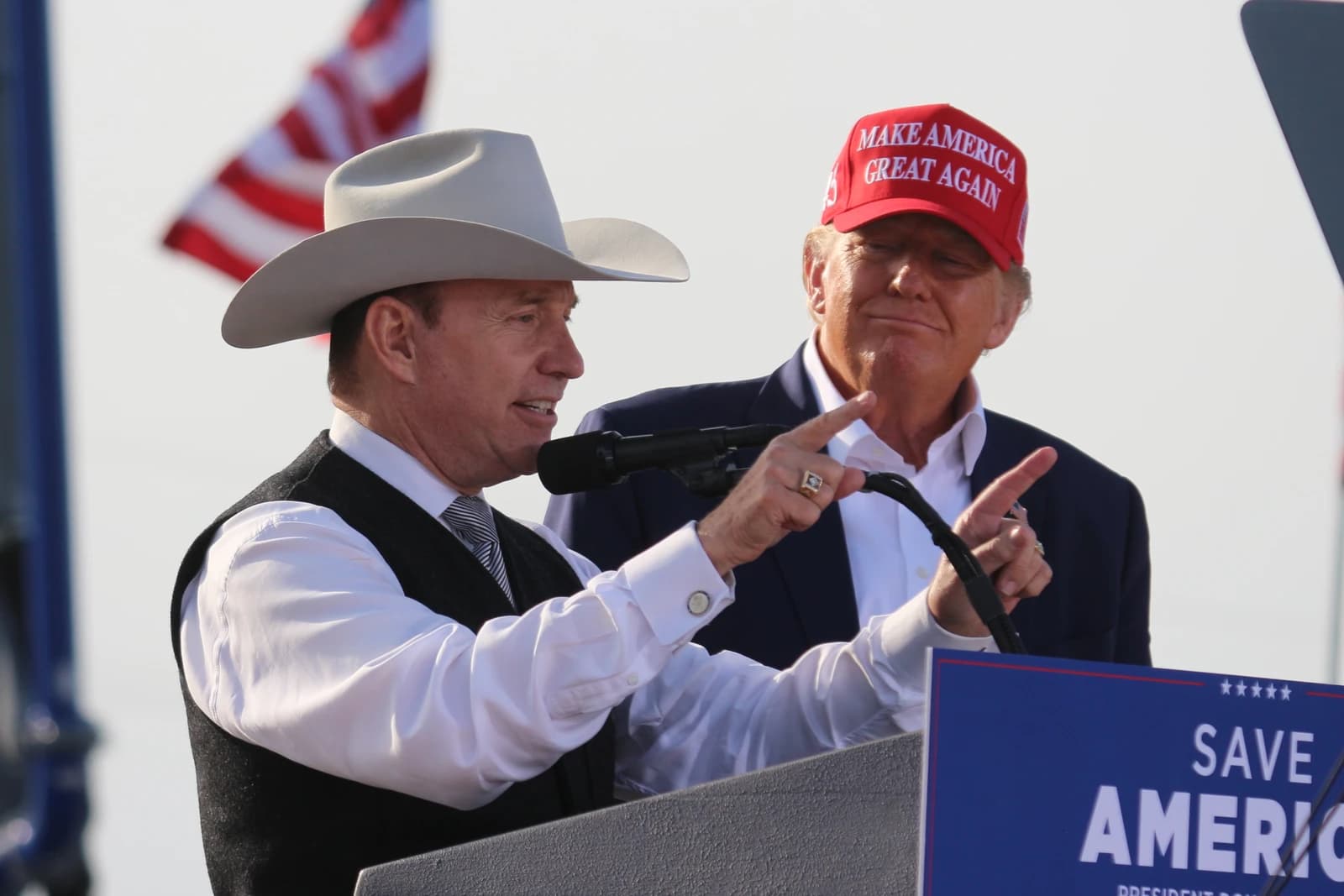 Charles Herbster, wearing a cowboy hat, speaks at a podium with Donald Trump standing behind him.
