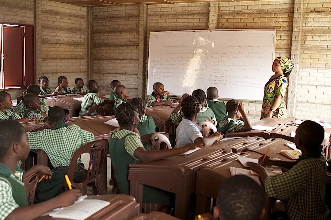 Students sit at desks in a classroom with a female teacher standing at a whiteboard.
