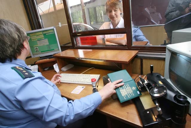 Border control officer in uniform at a desk, checking documents of a person through a window.