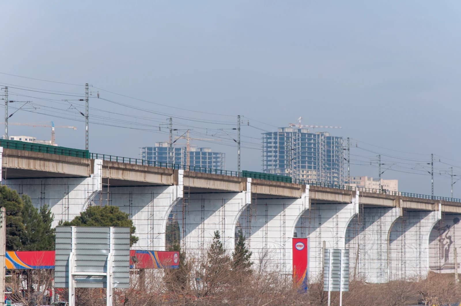 Railway bridge in Iran with electric lines, trees, and buildings visible.