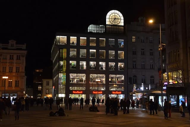 Exterior of Kotva Department Store in Prague at night, with illuminated windows and shoppers.