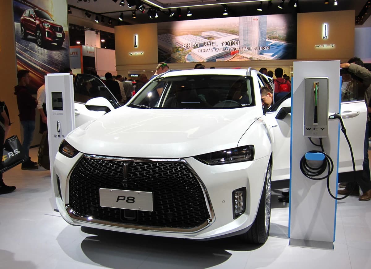 A white WEY P8 plug-in hybrid SUV is displayed on a show floor, with a charging station next to it.