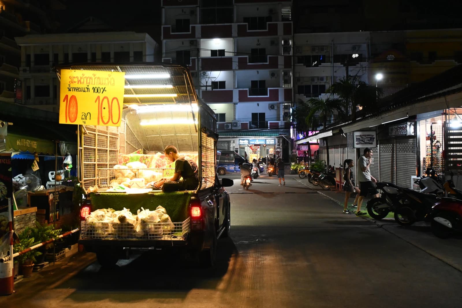 A street vendor sits in the back of a pickup truck selling produce at night on an urban street.