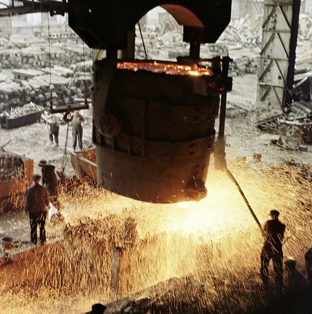 Workers perform teeming operation in a blast furnace plant, molten metal creating sparks.