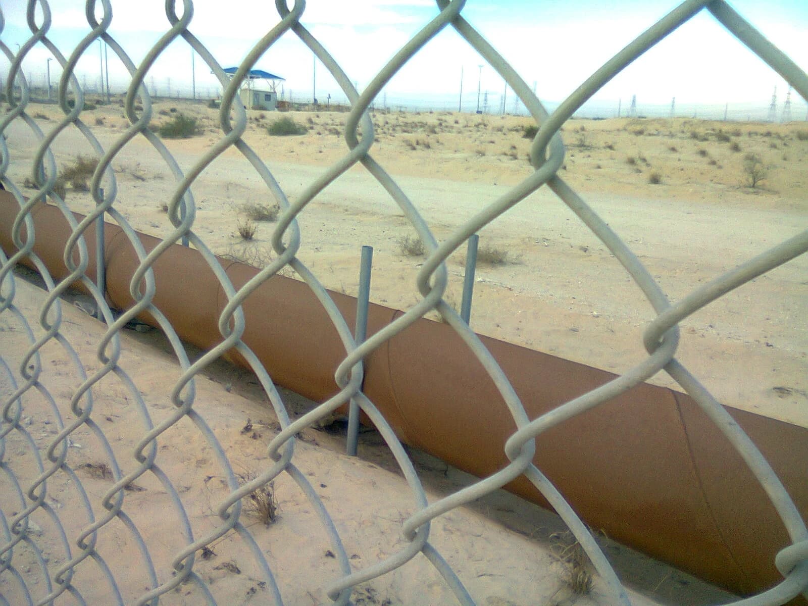 Oil pipelines crossing a desert landscape in Jubail, Saudi Arabia, viewed through a chain-link fence.