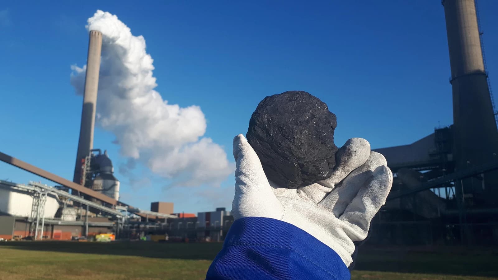 A gloved hand holds a lump of coal in front of a power plant with a smoking chimney.
