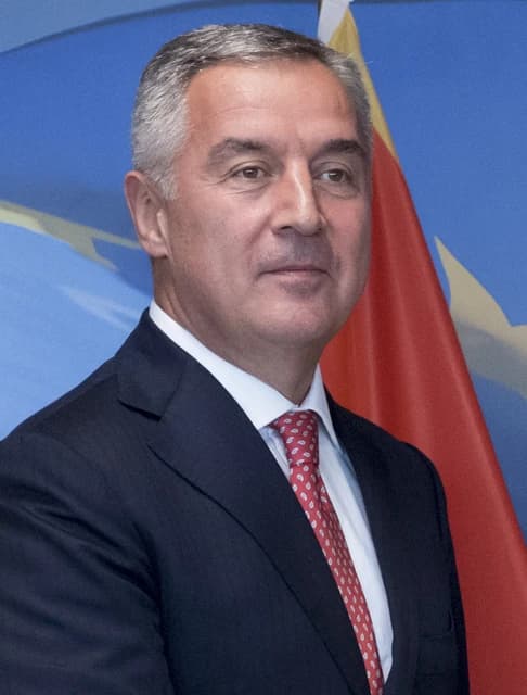 Milo Đukanović, President of Montenegro, stands in front of flags indoors.
